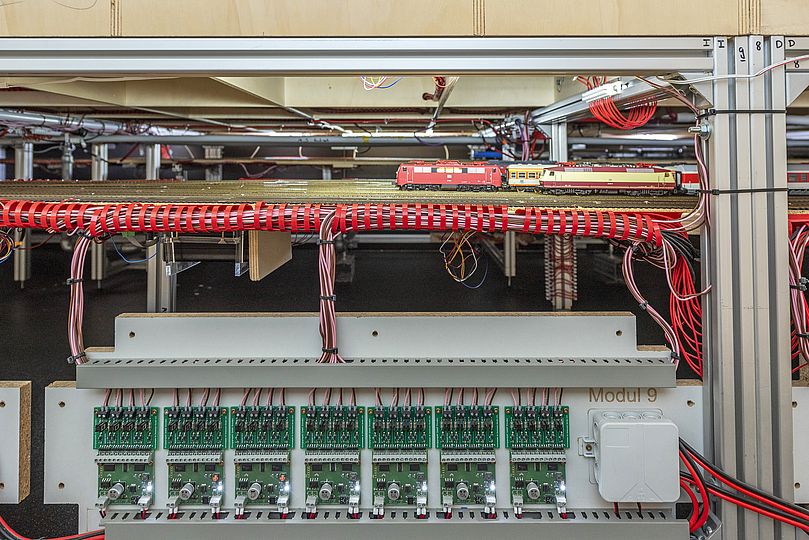 Von einem Brett mit Elektronikplatinen führen viele Kabel über Kabelbäumen zu den Gleisen der Modellbahn im Untergrund der Anlage.